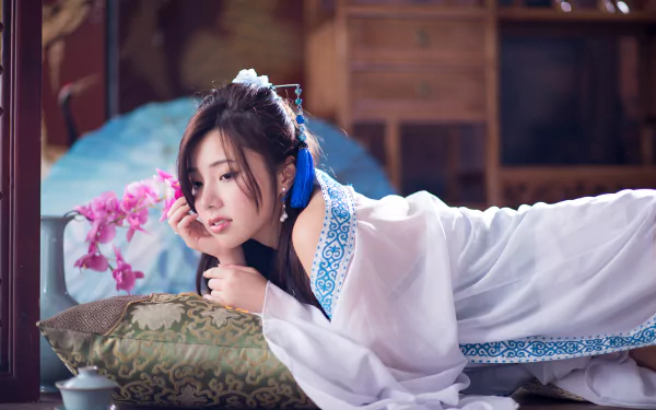 A woman in a flowing white dress rests on a cushion, surrounded by orchids and a vase, capturing an elegant Taiwanese aesthetic in this vibrant 4K Ultra HD wallpaper.