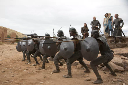 A dramatic scene from Game of Thrones featuring warriors with shields and spears in the foreground, while characters including Daenerys Targaryen and Missandei observe in the background.