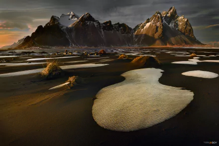 HD desktop wallpaper of a spring scene in Iceland featuring black sandy terrain interspersed with snow patches, under a dramatic sky with sharp mountain peaks in the background.
