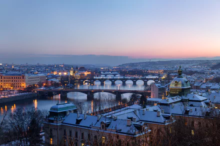 Winter view of Prague with snow-covered rooftops and multiple man-made bridges spanning the Vltava River at dusk, captured in 4K Ultra HD quality.