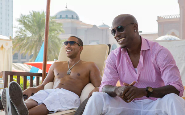Two characters from the movie Furious 7, Tej (Ludacris) and Roman Pearce (Tyrese Gibson), are sitting and smiling outdoors in a relaxed setting, with a luxurious backdrop in the image.