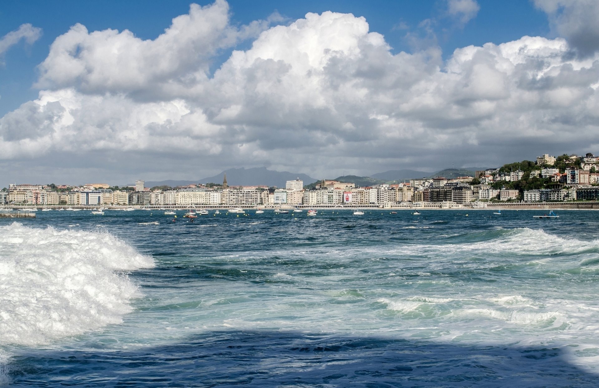Panoramic view of the coastal city of San Sebastián, Spain, featuring the shoreline, waves, and urban skyline under a partly cloudy sky, in HD quality.