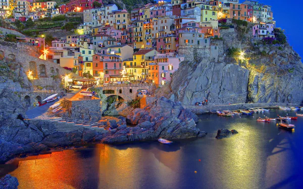 Vibrant night view of the man-made coastal village of Manarola, with colorful buildings perched on rocky cliffs reflecting in calm waters, captured in HD for a desktop wallpaper.