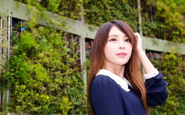 4K Ultra HD desktop wallpaper of a Taiwanese Asian model: a woman in a navy dress with a white collar posing by a leafy green fence, gazing softly toward the light.