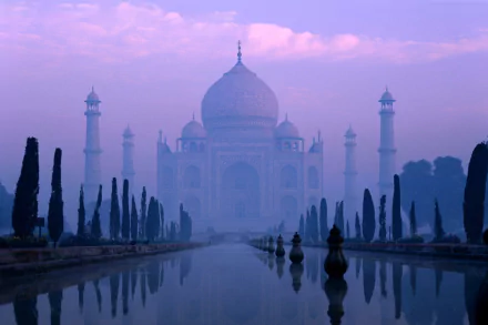 A serene view of the Taj Mahal at dawn, enveloped in mist, reflecting in the tranquil water with neatly trimmed trees, creating a stunning HD desktop wallpaper.