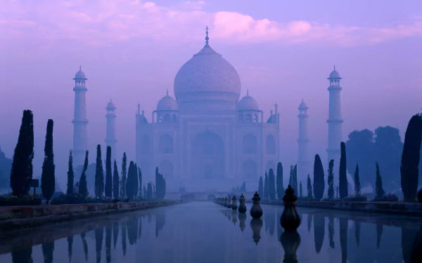 A serene view of the Taj Mahal at dawn, enveloped in mist, reflecting in the tranquil water with neatly trimmed trees, creating a stunning HD desktop wallpaper.