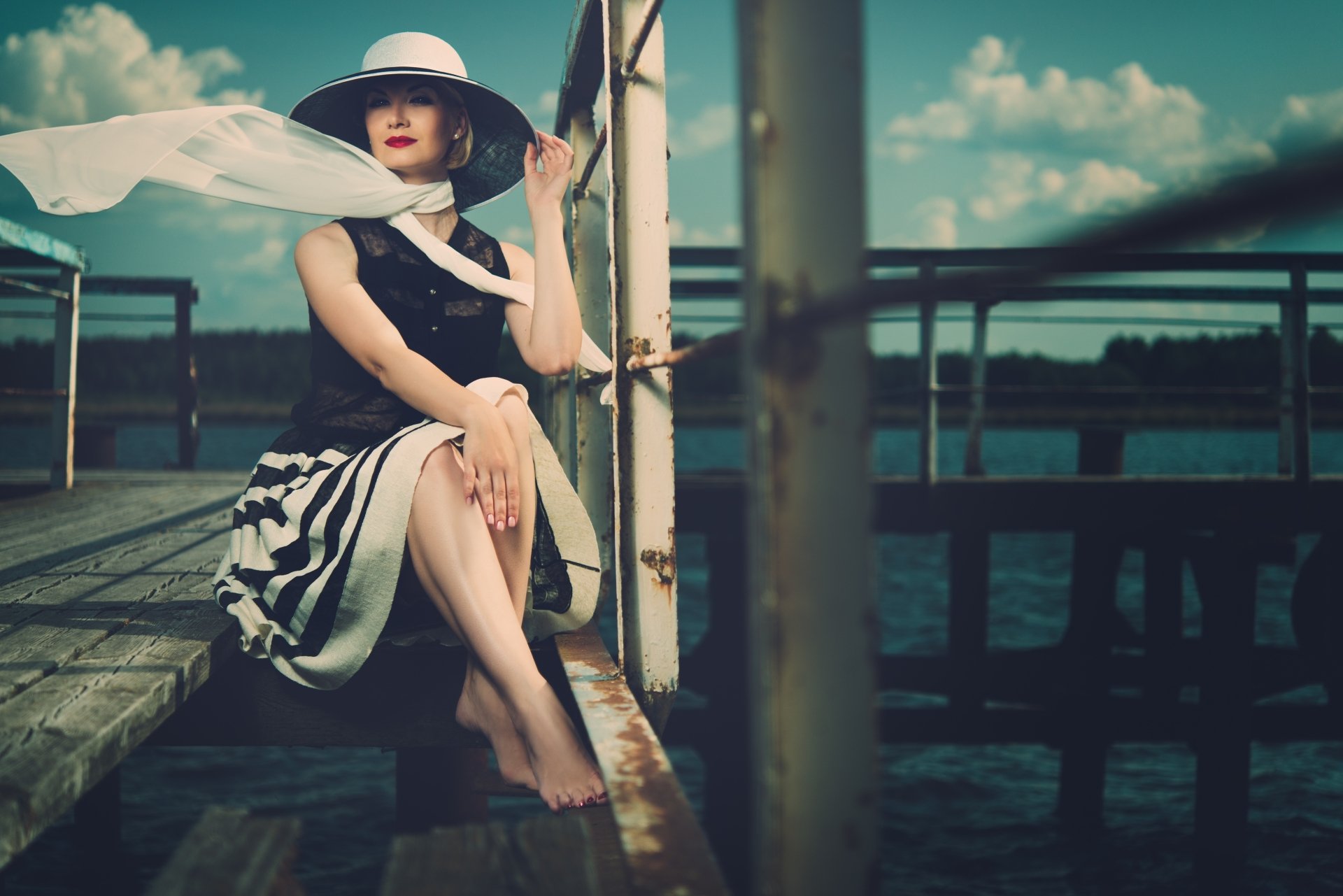 A vintage-style woman in a wide-brim hat and striped skirt sits barefoot on a dock against a serene sky, captured in 4K Ultra HD for PC desktop wallpaper.