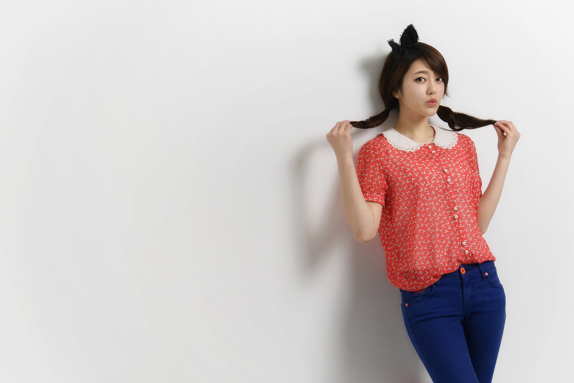 HD desktop wallpaper featuring an Asian woman with plaited hair and cat ears, wearing a red shirt with white polka dots and blue pants, posing against a plain white background.