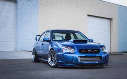 A blue Subaru Impreza parked against a white industrial building, showcased as a high-definition PC desktop wallpaper and background.
