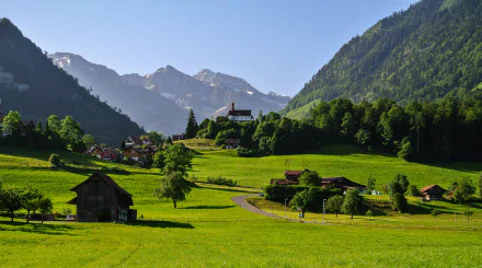 HD landscape wallpaper of a Swiss village in a meadow and valley, featuring a church, surrounded by mountains. It's a serene morning scene epitomizing the tranquil beauty of Switzerland.