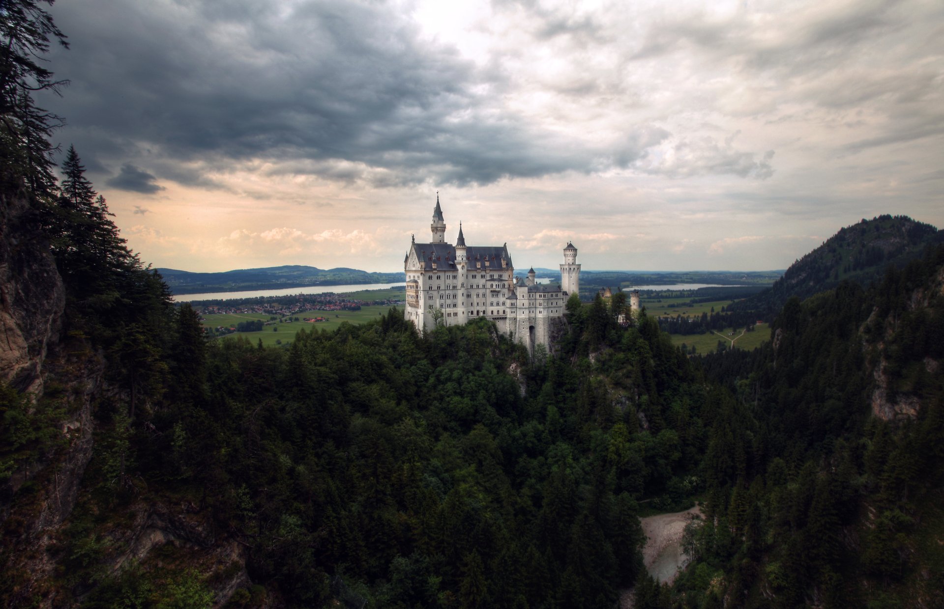 Neuschwanstein Castle perched above a Bavarian forest under dramatic clouds, panoramic landscape view — 5K Ultra HD PC desktop wallpaper.