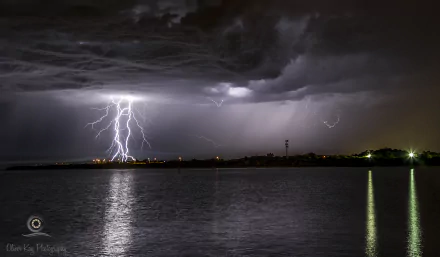 Night lightning photography: a dramatic bolt lights the sky over a shoreline, reflections rippling on the water — 2K Quad HD PC desktop wallpaper background.