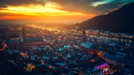 HD PC desktop wallpaper: man-made Heidelberg cityscape at sunset, glowing riverside buildings and rooftops beneath a dramatic sky.