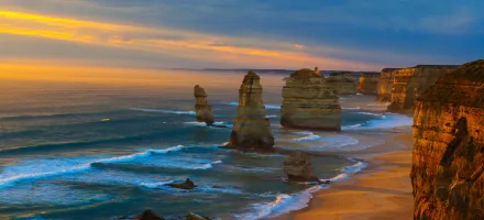 Sunset over the limestone stacks of The Twelve Apostles along the Victoria coastline in Australia, with waves gently washing the shore under a colorful sky.