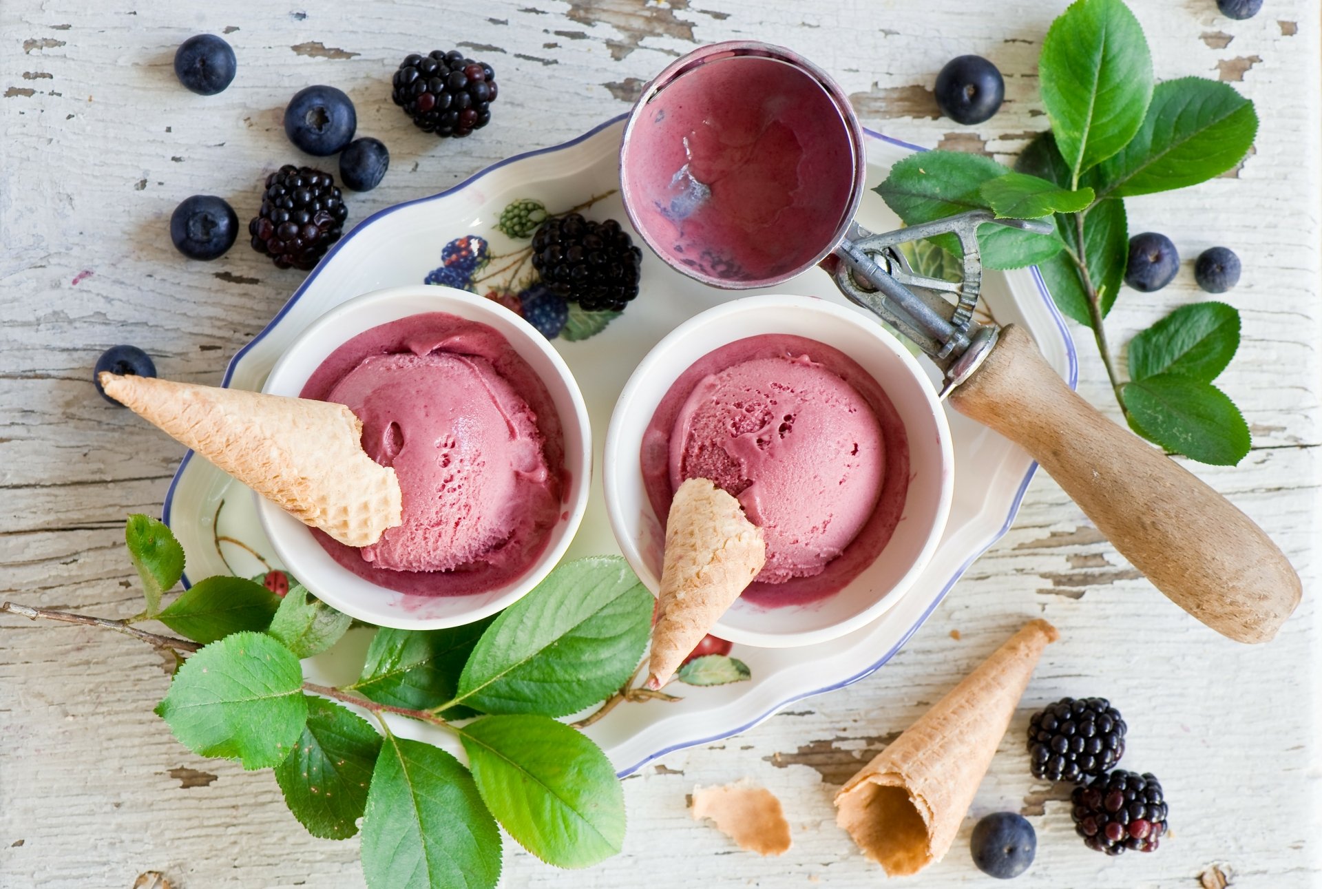 A 4K Ultra HD still life image featuring two bowls of berry ice cream with cones, fresh blueberries, blackberries, and green leaves on a rustic wooden surface.