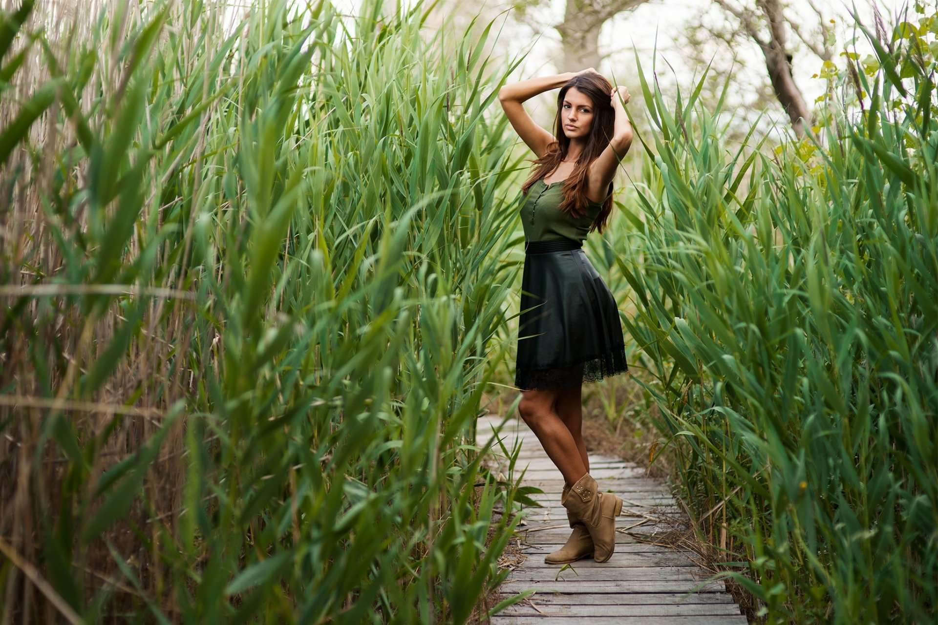 A brunette woman in boots stands on a boardwalk surrounded by lush plants, striking a model pose. HD desktop wallpaper and background.