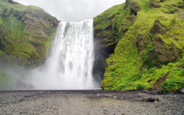 Skógafoss waterfall in Iceland cascading between mossy green cliffs, mist rising over a rocky shoreline — 4K Ultra HD PC desktop wallpaper and nature background.