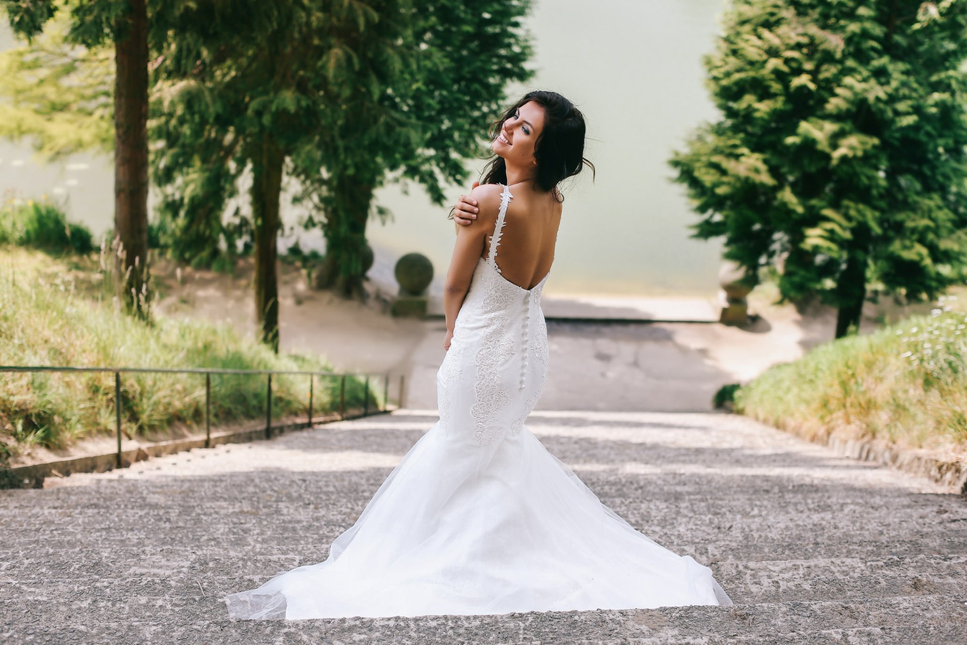 A brunette bride models an elegant wedding dress, posing gracefully on a stone pathway surrounded by greenery. This HD image captures a beautiful, serene moment in nature.