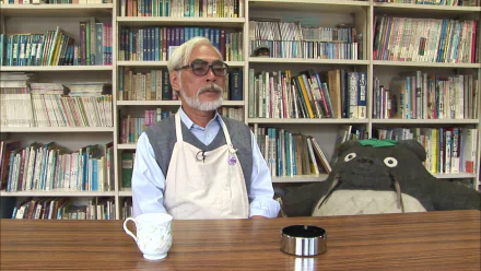 HD desktop wallpaper featuring a man at a desk with a Totoro plushie, reflecting themes associated with Hayao Miyazaki.