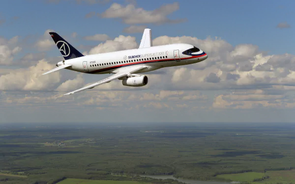 HD desktop wallpaper featuring a Sukhoi SuperJet 100 airplane jet flying above a green landscape under a partly cloudy sky.