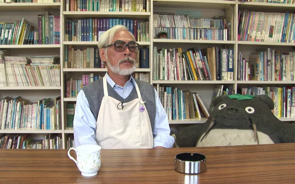 HD desktop wallpaper featuring a man at a desk with a Totoro plushie, reflecting themes associated with Hayao Miyazaki.