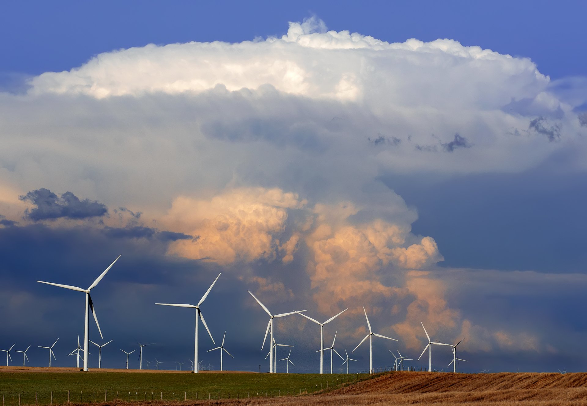 HD desktop wallpaper of man-made wind turbines on a Colorado plain beneath a glowing cumulus thunderhead cloud at sunset.