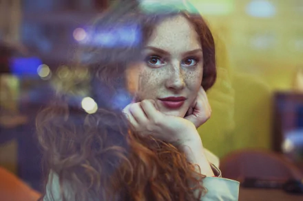 HD desktop wallpaper of a freckled redhead woman/model with brown eyes, resting her chin on her hands and gazing thoughtfully through a window.