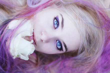A striking HD wallpaper featuring a model with blonde hair and blue eyes, softly framed by pink and purple hues, holding a white rose close to her face.