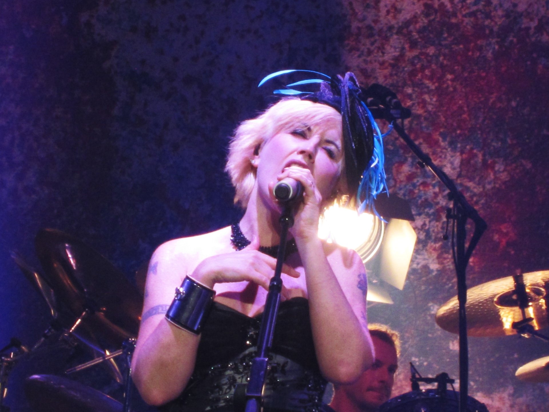 Female singer performing onstage, gripping a microphone with drums behind, vivid colored lighting and textured backdrop — 4K Ultra HD PC desktop music wallpaper.