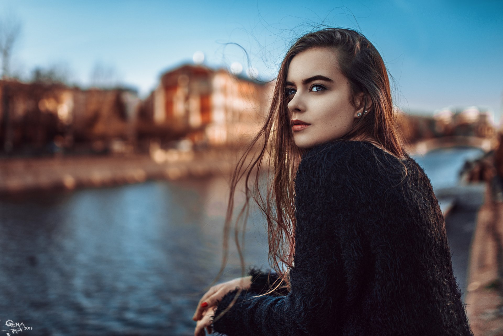A brunette woman with blue eyes gazes thoughtfully to her side, set against a serene backdrop of water and city buildings, creating an inviting HD desktop wallpaper.