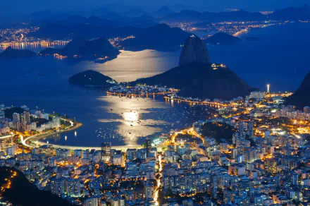 A stunning HD wallpaper of Rio de Janeiro at night, showcasing vibrant city lights along the coastline, with Sugarloaf Mountain prominently rising against a serene bay.