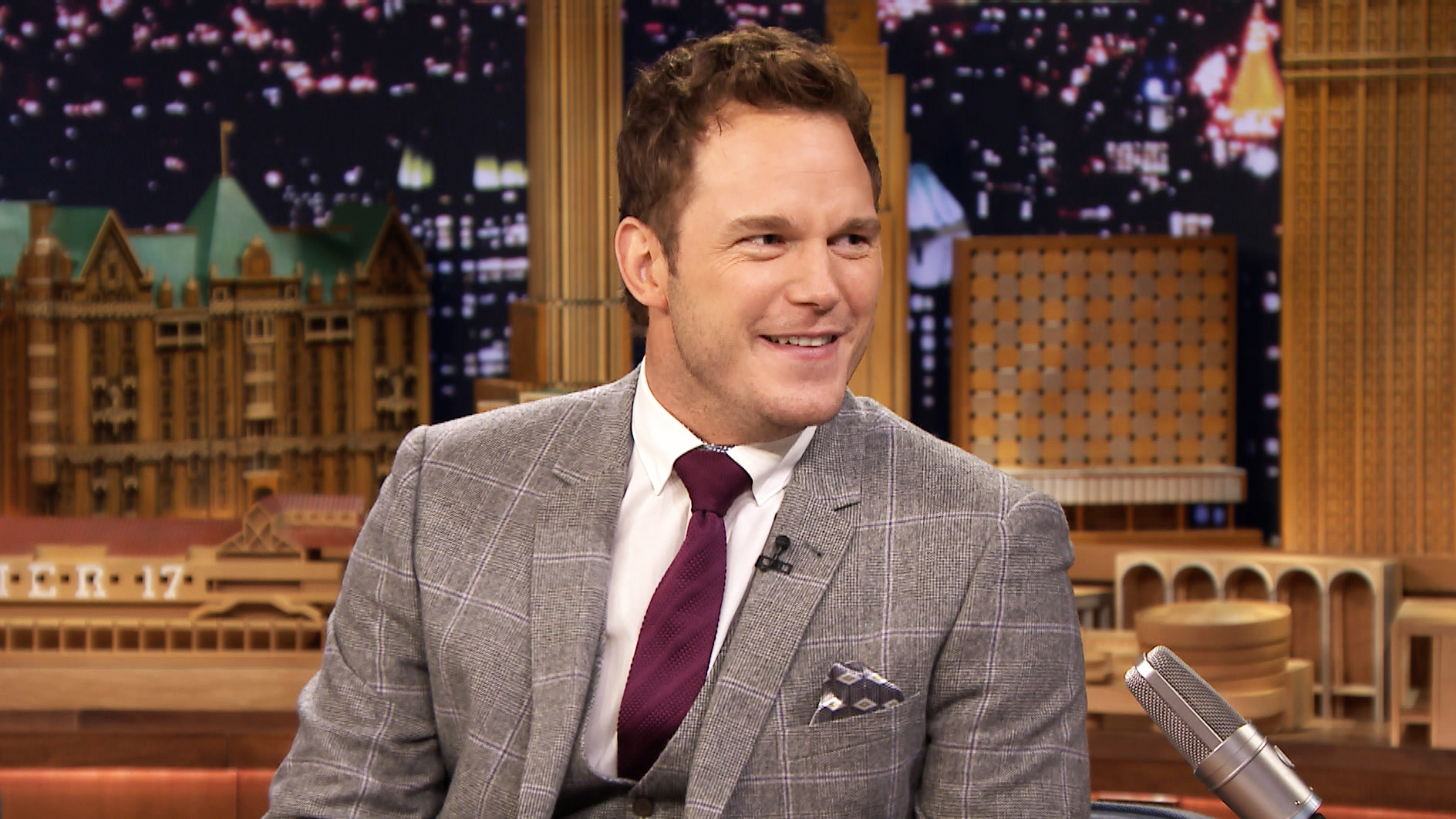 HD desktop wallpaper of Chris Pratt wearing a grey suit with a maroon tie, smiling during a TV show interview, with a cityscape background.