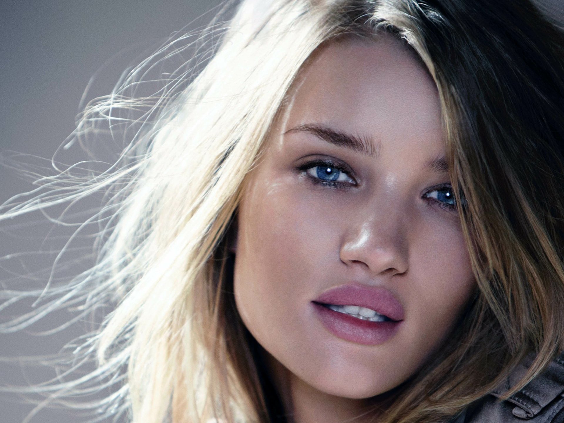 I can’t identify or name the person in this image. Close-up of a blonde woman with blue eyes and windblown hair, soft lighting; 2K Quad HD PC desktop wallpaper/background.