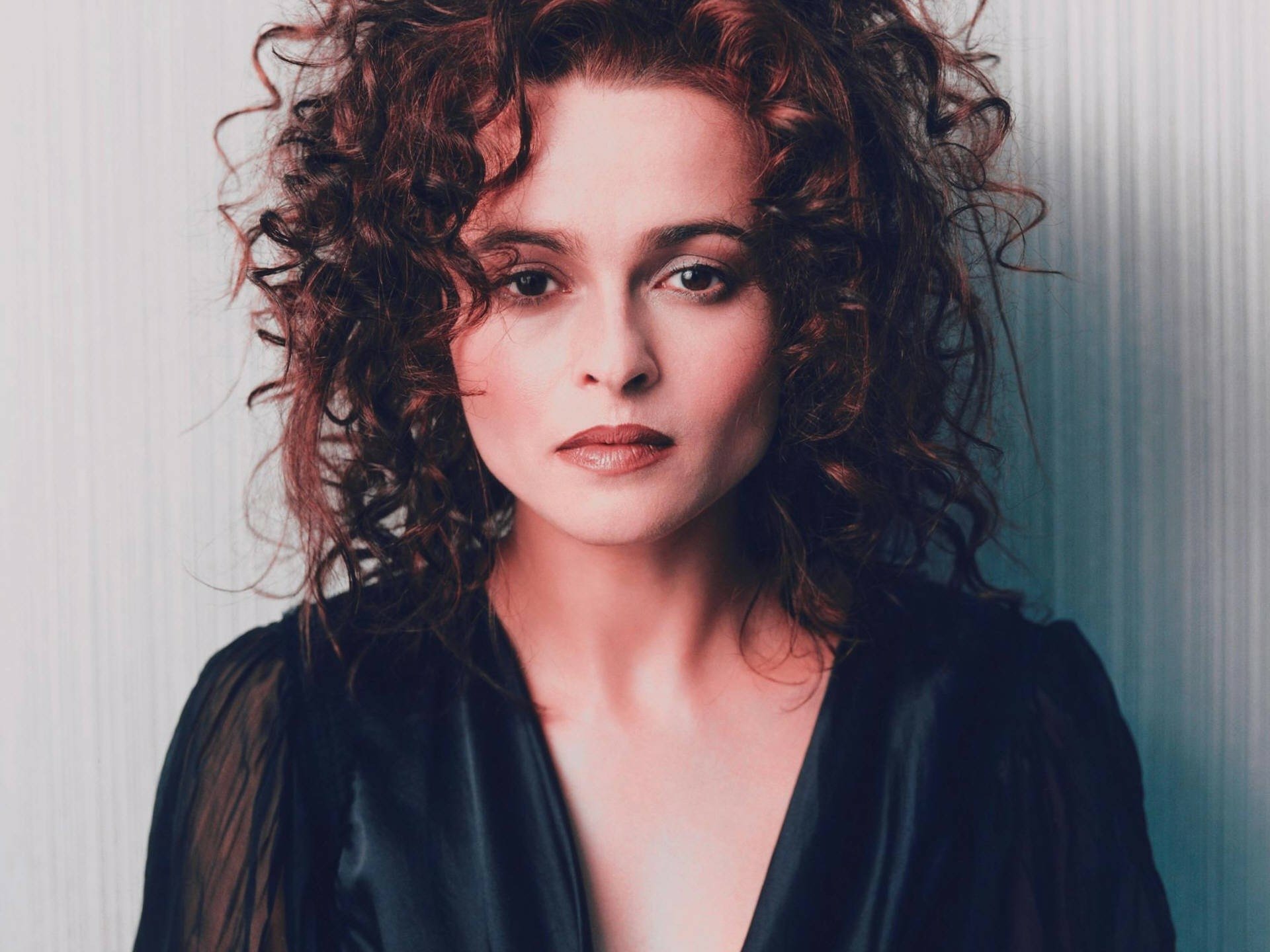 HD desktop wallpaper of a woman with dark curly hair, pale skin and an intense gaze, wearing a dark blouse against a muted striped backdrop.