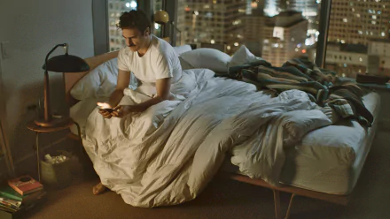 Joaquin Phoenix in a scene from the movie Her, sitting on a bed in a softly lit room with a cityscape visible through the window, HD PC desktop wallpaper.