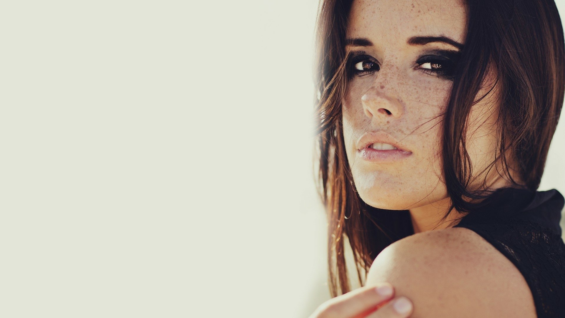 HD PC desktop wallpaper: close-up portrait of a woman with dark hair and light freckles looking over her shoulder against a pale, minimalist background.
