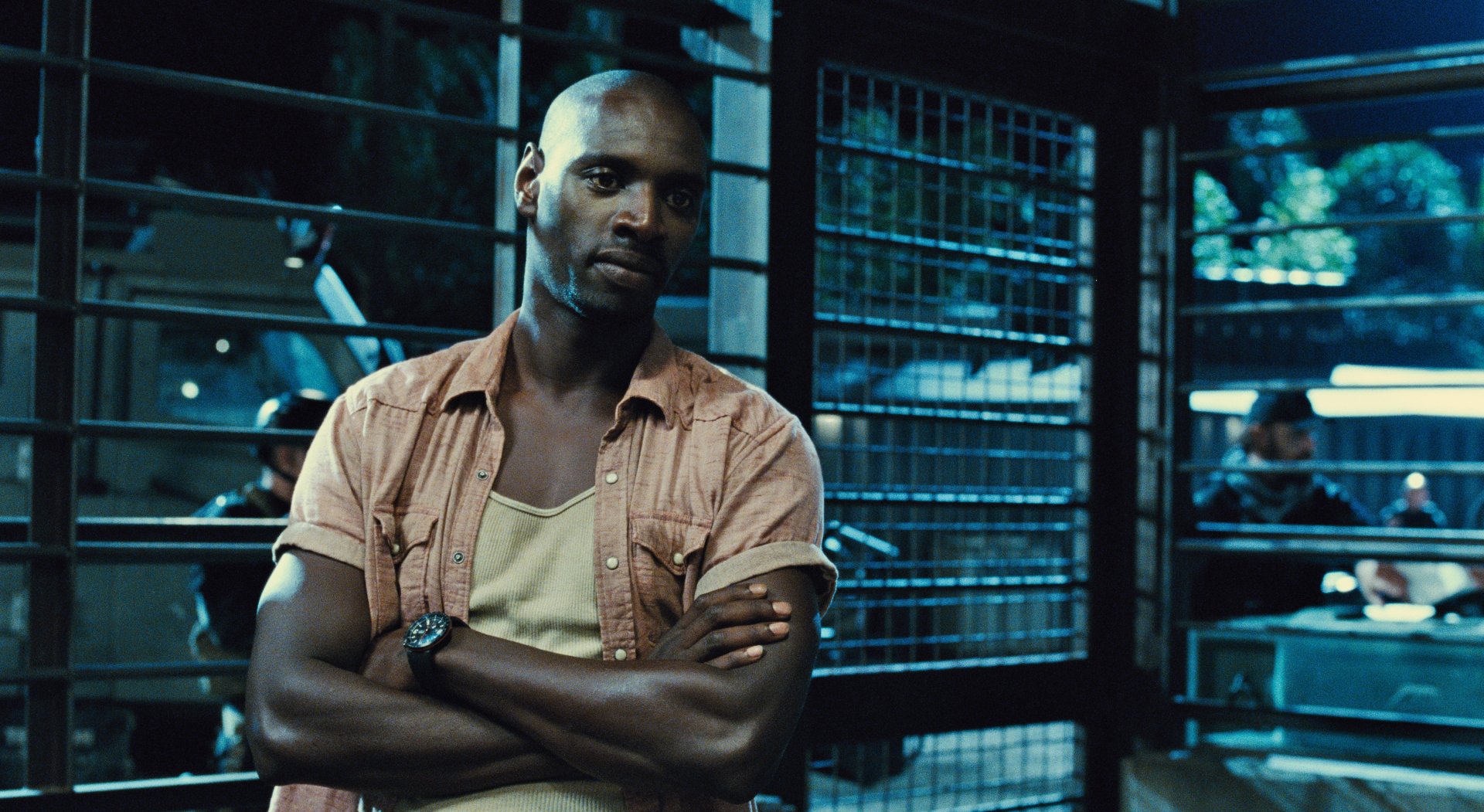 HD PC desktop wallpaper featuring Omar Sy in a scene from the movie Jurassic World, standing with arms crossed in a dimly lit, industrial setting.