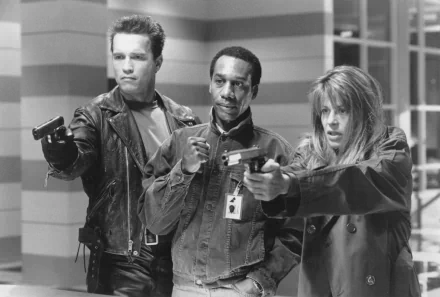 A black and white image from Terminator 2: Judgment Day featuring Arnold Schwarzenegger as the Terminator, Joe Morton as Miles Dyson, and Linda Hamilton as Sarah Connor, all wielding guns.