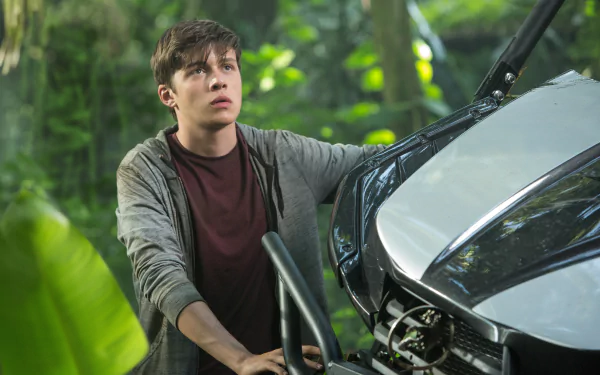 A young man, Nick Robinson, appears concerned while inspecting a vehicle in a lush jungle setting, capturing a tense moment from the movie Jurassic World in stunning 4K Ultra HD.