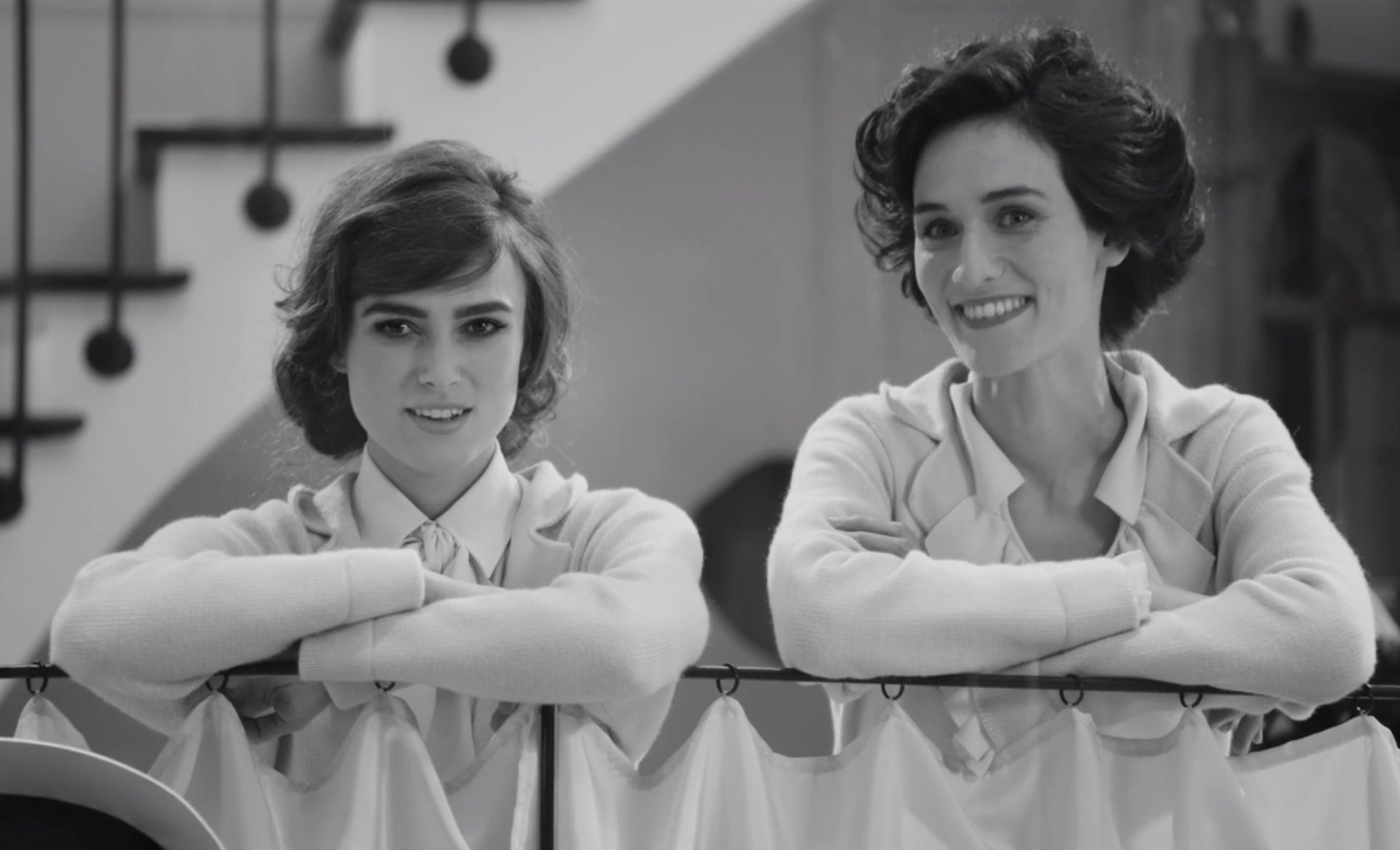 Black-and-white HD PC desktop wallpaper film still (2017): two young women in period clothing smiling and leaning on a railing inside a house.