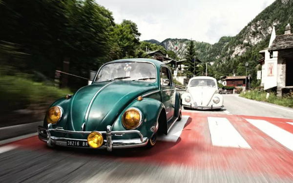HD PC desktop wallpaper featuring two classic Volkswagen Beetle vehicles driving on a scenic mountain road surrounded by greenery and rustic buildings.