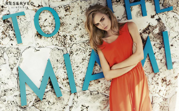 A blonde model poses in an orange dress against a textured wall featuring bold blue letters spelling TO THE MIAMI, capturing a stylish and vibrant atmosphere.