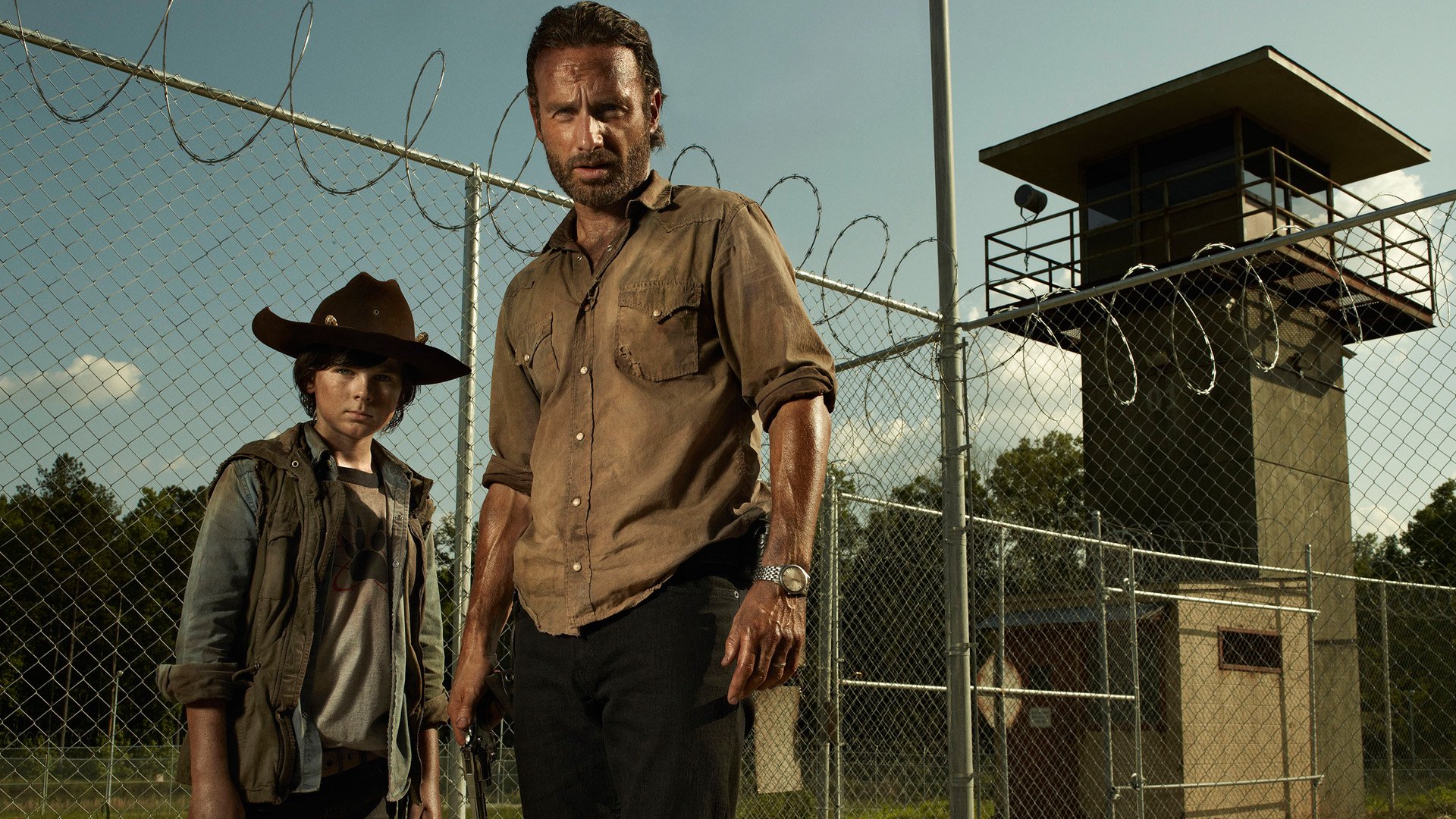 Rick Grimes and Carl Grimes stand in front of a prison fence, showcasing a tense moment from the TV show The Walking Dead. An intense atmosphere reflects their survival struggle.