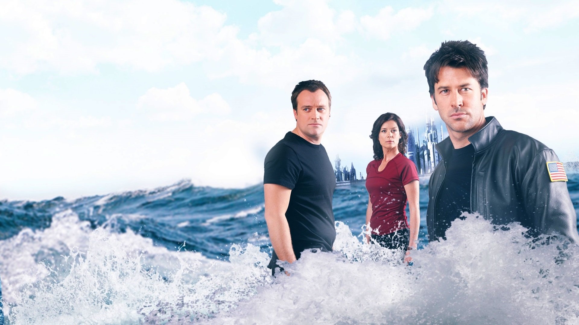 HD PC desktop wallpaper of the TV show Stargate Atlantis: three characters standing amid crashing ocean waves with a distant city skyline under a cloudy sky.