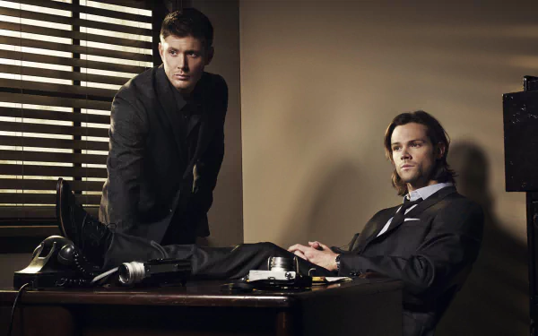 Two characters from the TV show Supernatural are seated at a desk, one standing beside it. The scene captures a tense moment, with dramatic lighting enhancing the atmosphere.