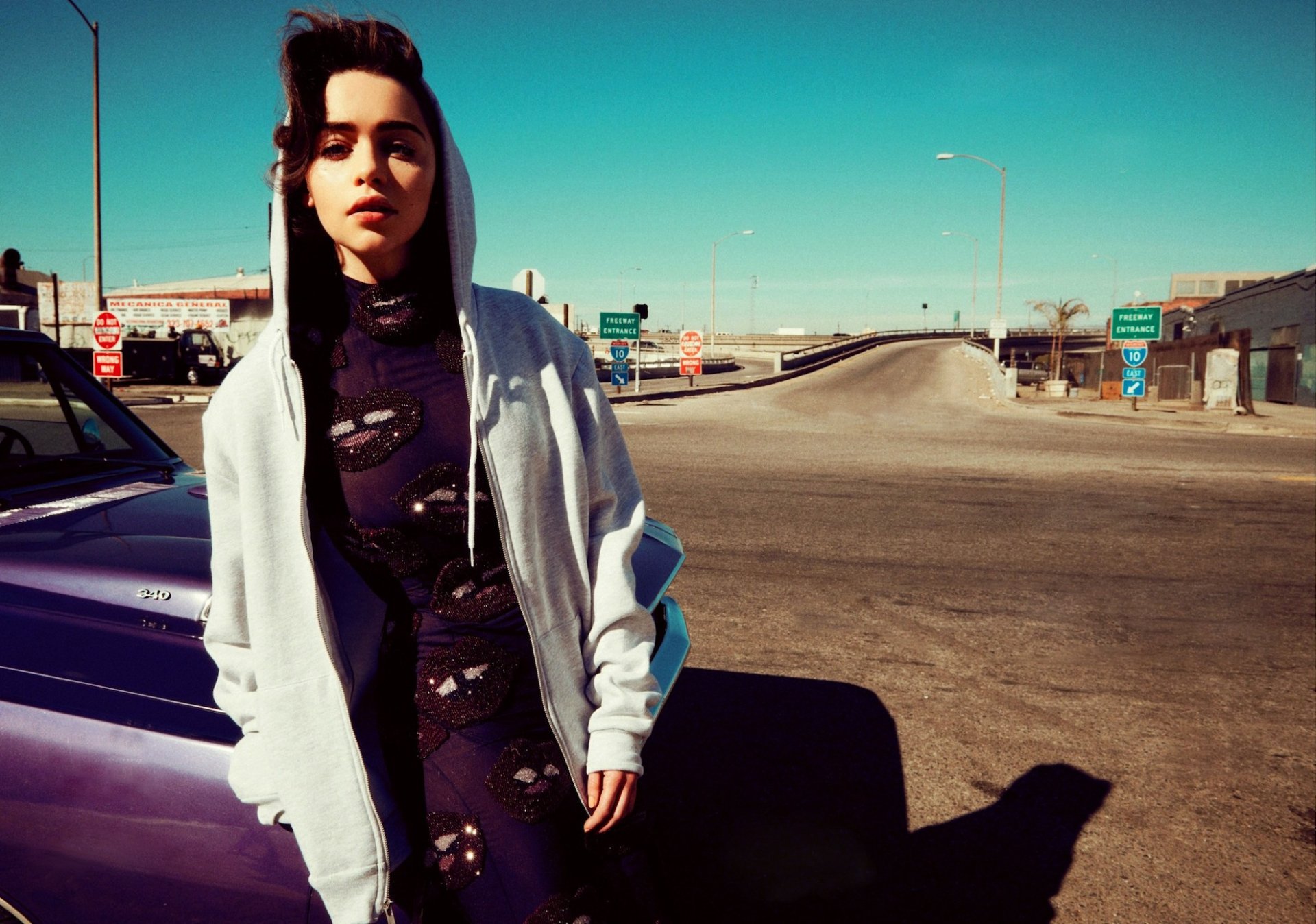 Brunette actress Emilia Clarke poses confidently in a stylish hoodie against a vibrant outdoor backdrop, capturing a striking moment in this high-definition wallpaper.