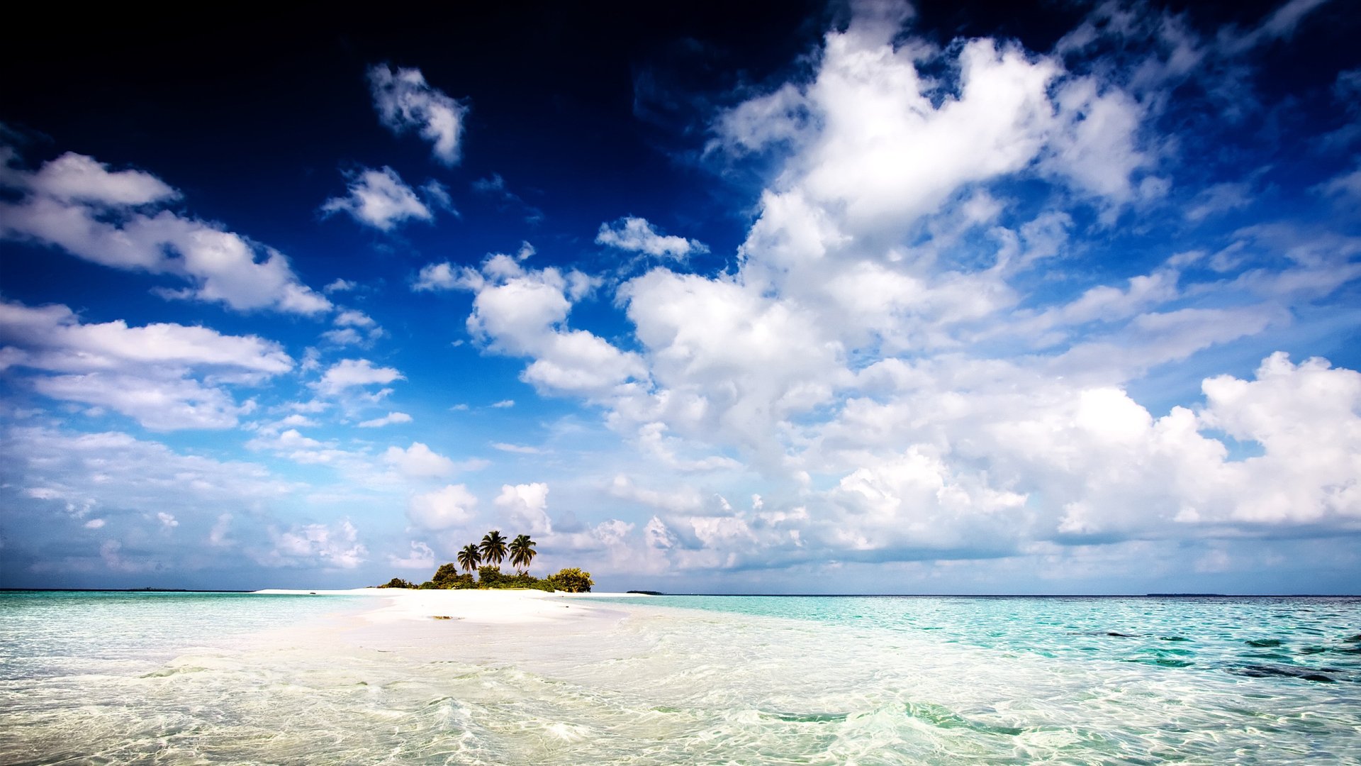 HD PC desktop wallpaper of a serene beach with clear turquoise water, a small island of palm trees, and a vibrant sky filled with scattered clouds.