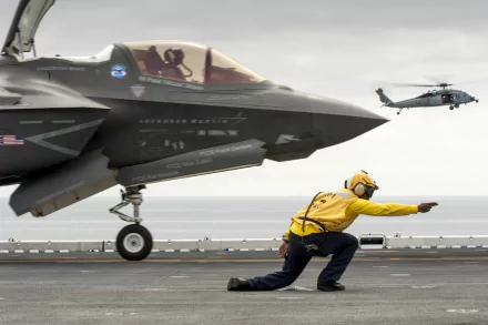Military scene: Lockheed Martin F-35 Lightning II on an aircraft carrier deck, a yellow-jacketed deck crew member directing operations with a helicopter in the distance — 2K Quad HD PC wallpaper.