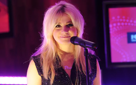 2K Quad HD PC desktop wallpaper of a blonde pop singer smiling into a microphone on a pink-purple lit stage, music-themed background.