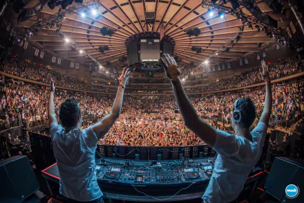 HD PC desktop wallpaper: high-energy music scene showing two DJs at the booth raising their arms to a packed, illuminated arena crowd under stadium lights.